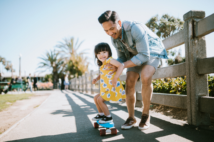 A dad helps his little girl go skateboarding, holding her waist for support. Shot in Los Angeles, California by the Santa Monica Pier.