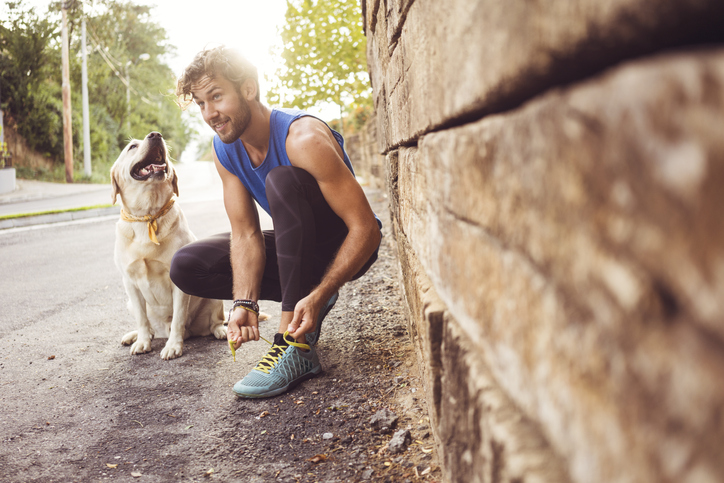 Young man jogging out with his dog, stopping on the side of the road to tie his shoe