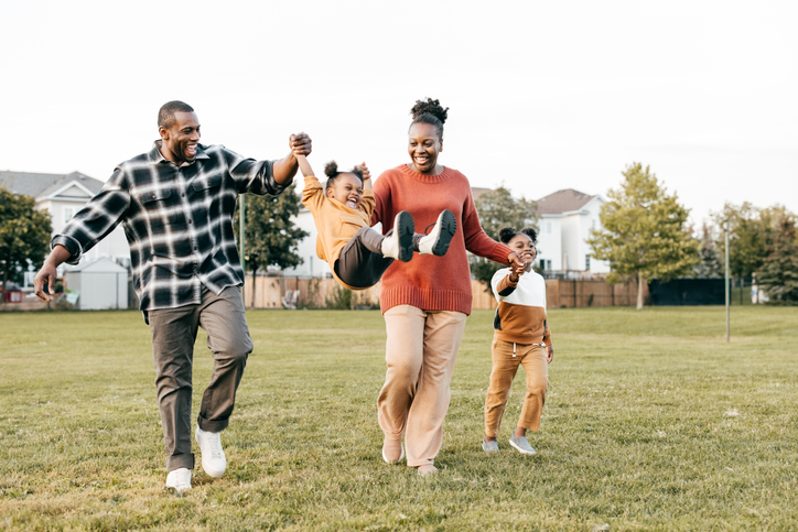 Family playing in the grass outside. Parents are swinging one child in the air between them and another child walking beside them, holding her mother's hand