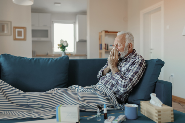 Cold exhausted senior man with flu wrapped in a warm blanket blowing his nose with a tissue in the livingroom