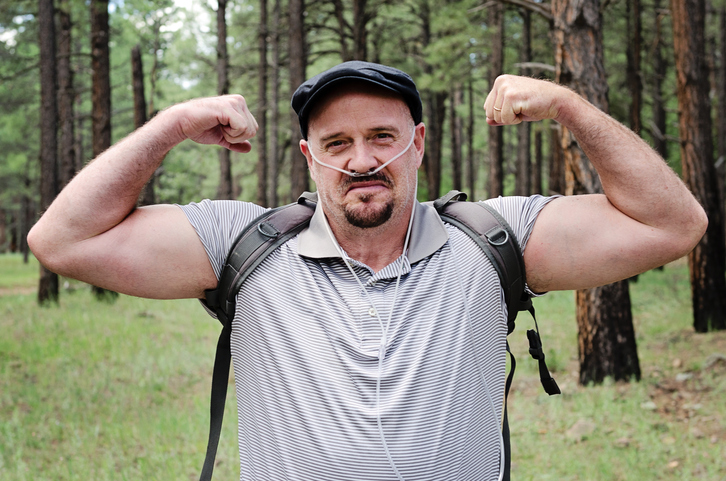 Middle aged man flexes his muscles as he hikes in the woods wearing therapeutic oxygen