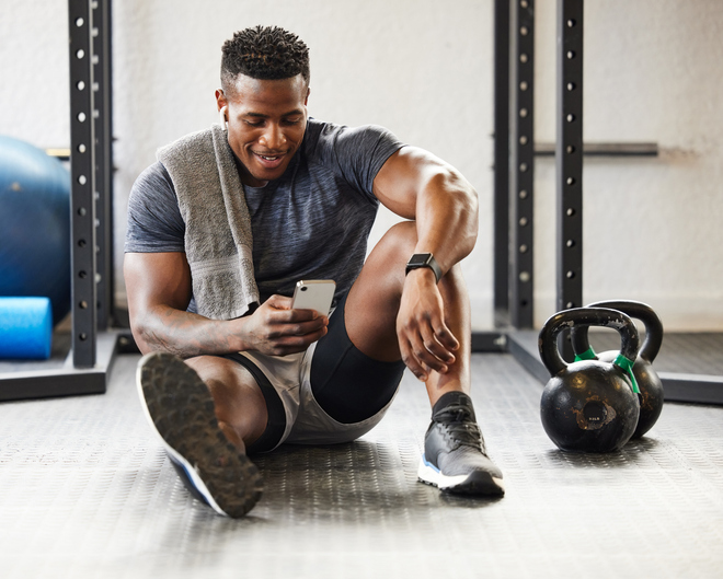 man sitting on the floor in a gym, with weights beside him, checking his health app on his phone