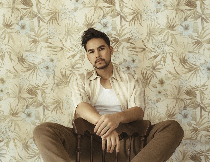 Tyler Shaw sits cross legged in front of a wallpapered wall.