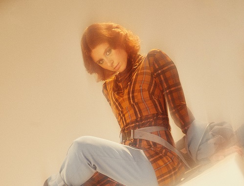 Kiesza sits on the ground wearing jeans and an orange top.