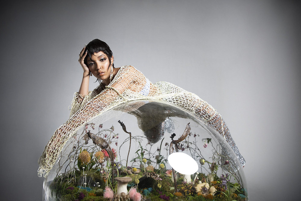 Tinashe lays on top of a clear globe filled with an eclectic mix of plant life.