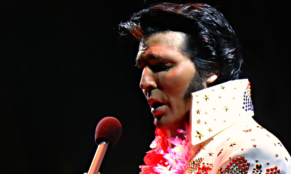 An Elvis impressionist sings into a microphone at a concert.