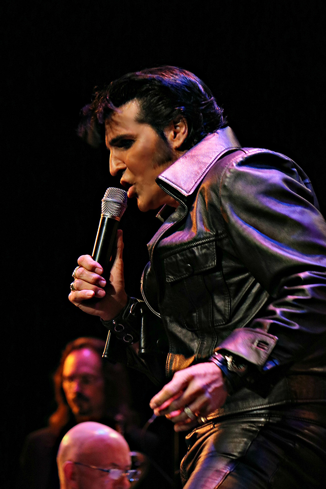 An Elvis impressionist sings into a microphone at a concert.