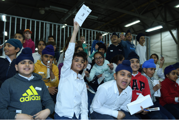 Kids sit in a crowd in bleachers and hold up pieces of paper as they bid on items in an auction