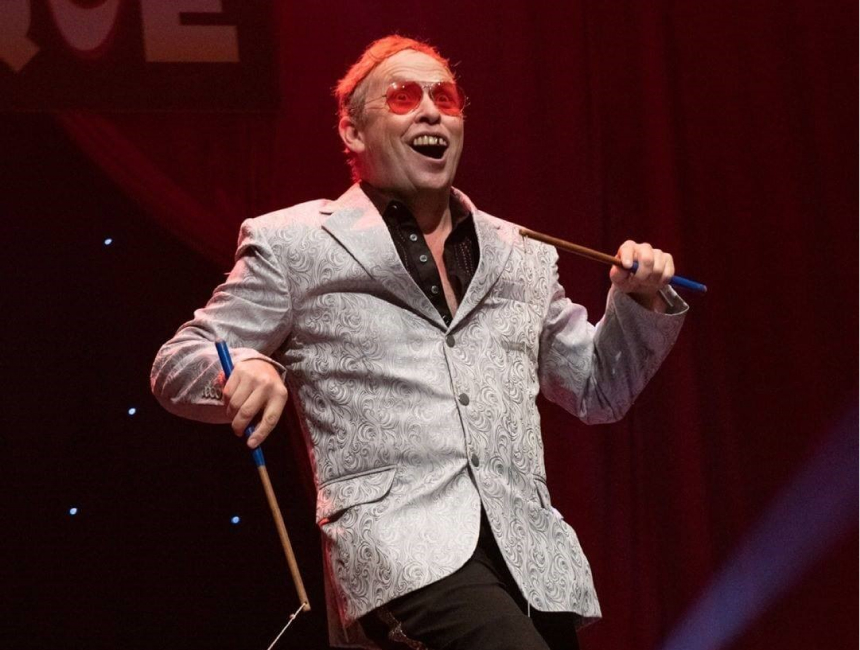 Vinyl Burns performs on stage wearing a grey suit jacket and sunglasses with red lenses