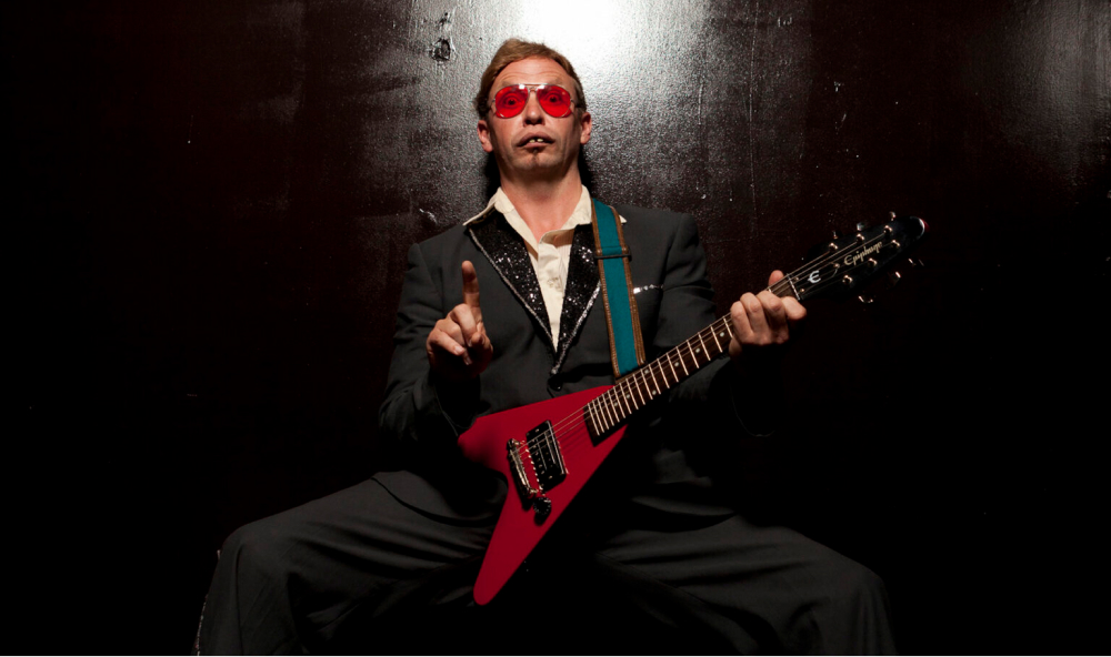 Vinyl Burns sits with red guitar and sunglasses facing camera