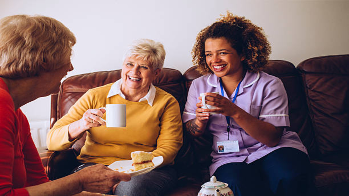 Two elderly women sat on the couch having tea with female caregiver, smiling.