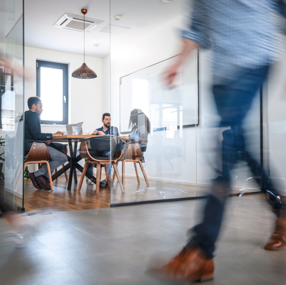 Person walking in front of a meeting room with people sitting around a table.