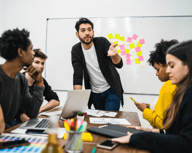 5 people at a conference table. One man is standing with sticky notes behind him.