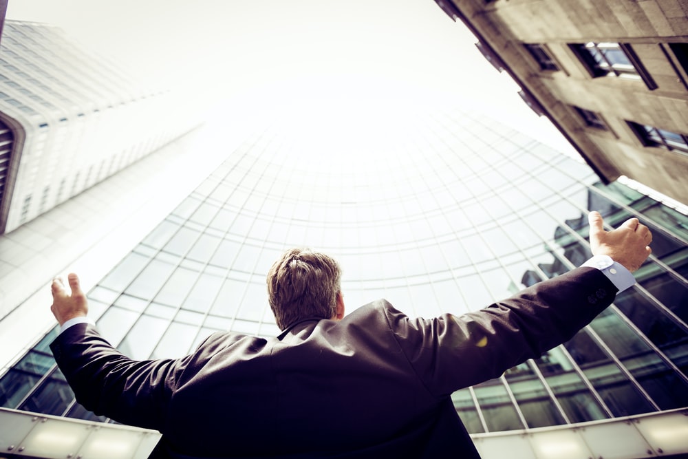 A business man outside of a tall building with his arms up to the sky