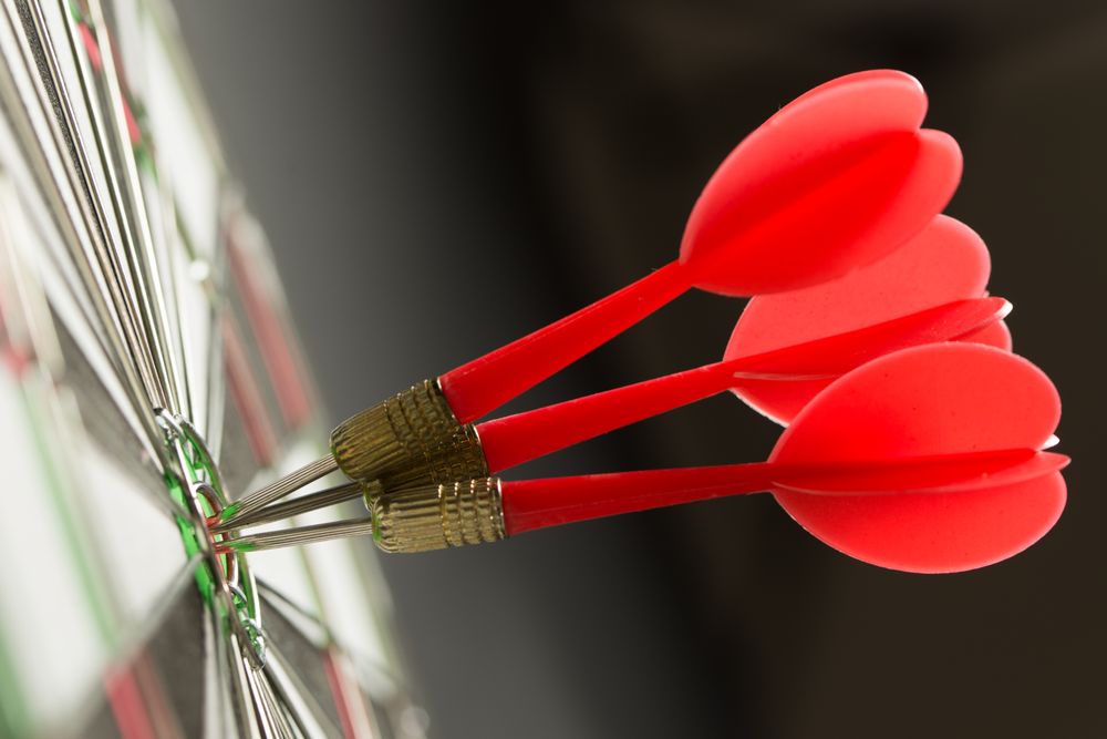 Close up image of 3 darts hitting the bullseye of the same dark board.