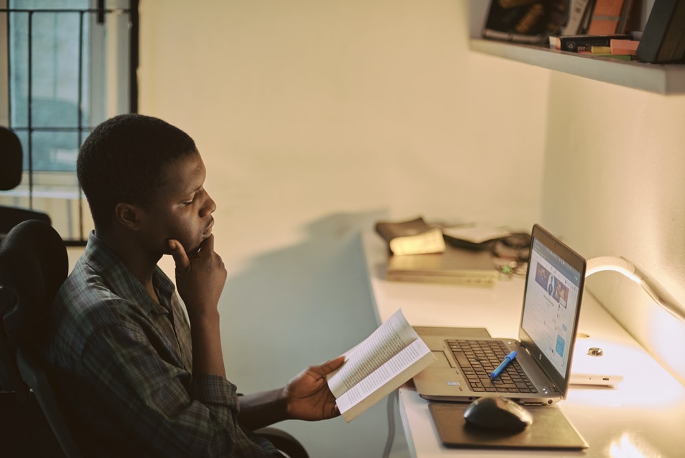 a man in front of a computer, reading a book.
