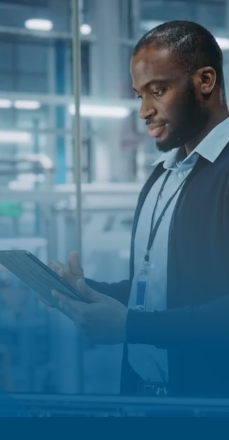 A man in a glass office overlooking an industrial work environment looking at a tablet computer