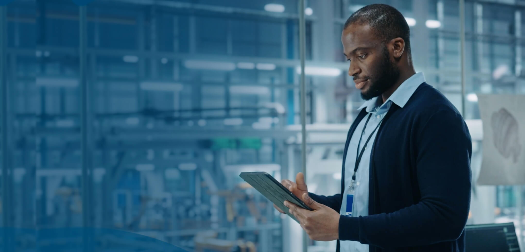 A person in a glass office overlooking an industrial work environment looking at a tablet computer