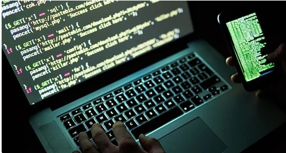 A person sitting in front of a laptop, attempting to hack into a system while holding a cell phone.