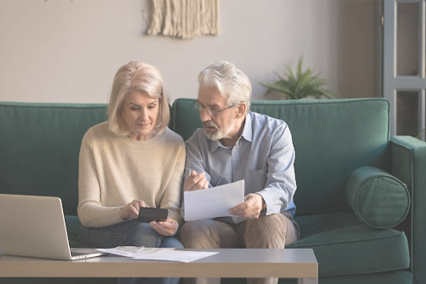 An elderly couple pre-planning their funeral; sitting on a couch while holding papers and a phone with a laptop on a coffee tabl