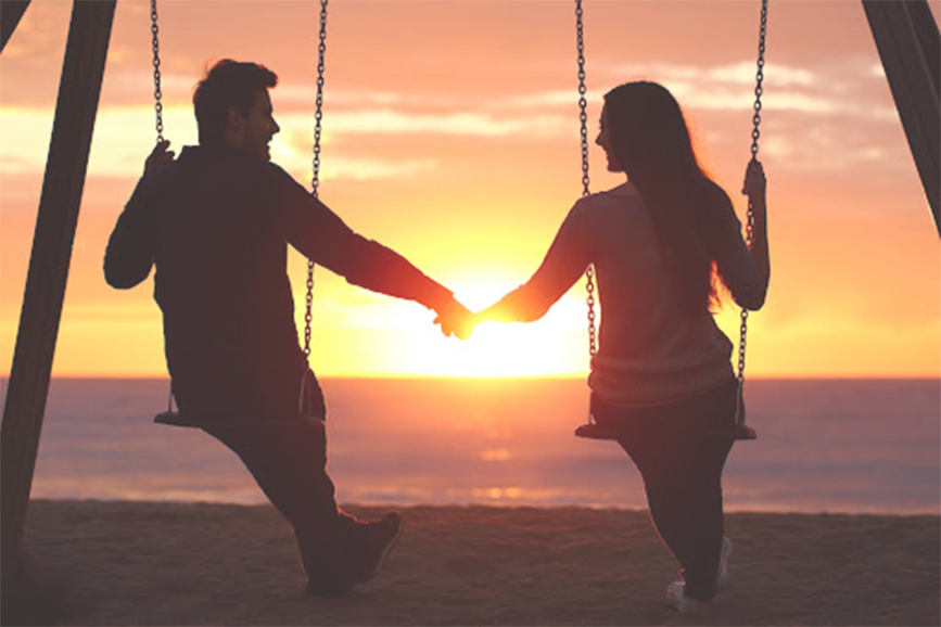 A smiling couple holding hands on swings at the beach while peacefully watching the sunset
