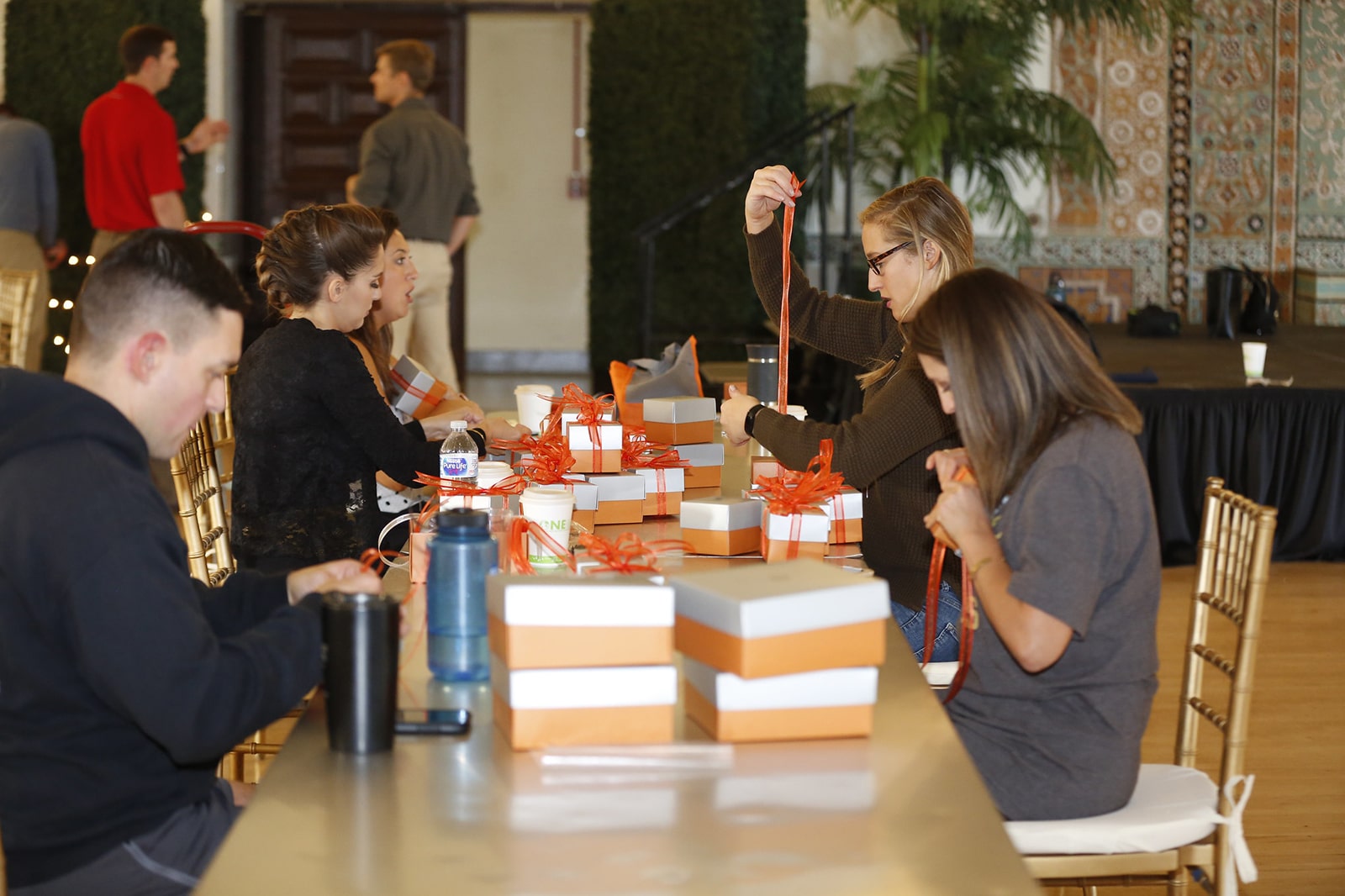 Five people sitting at a table, there are gift boxes on the table and each person is working on putting red ribbons on the gift boxes. The boxes are copper colored with a white lid.