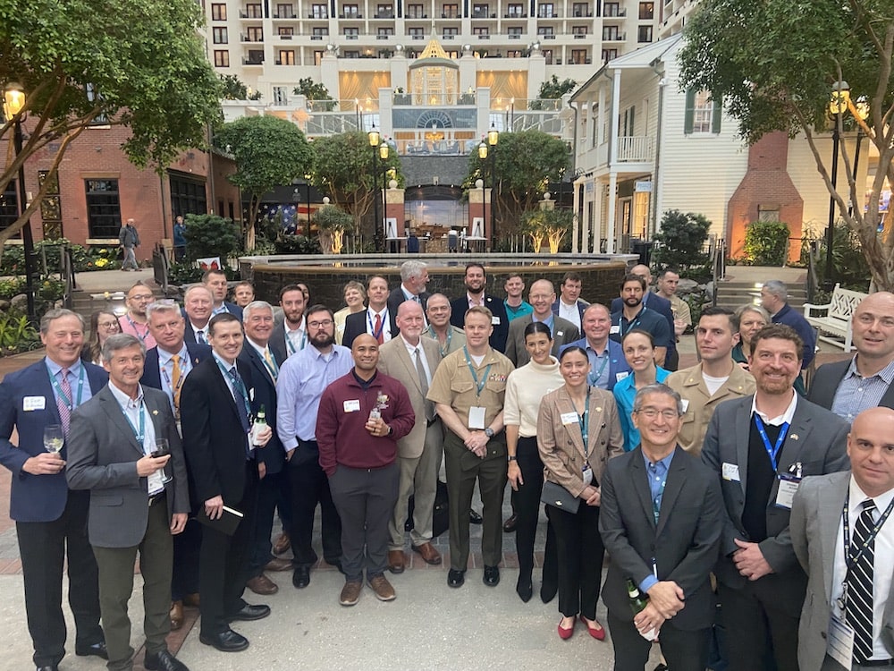 Group of approximately forty people all standing in a half circle and facing the camera. They all have lanyards on, many have drinks, some are in suits and others are wearing military uniforms. There is a large building behind the group as well as a circular man made pond.