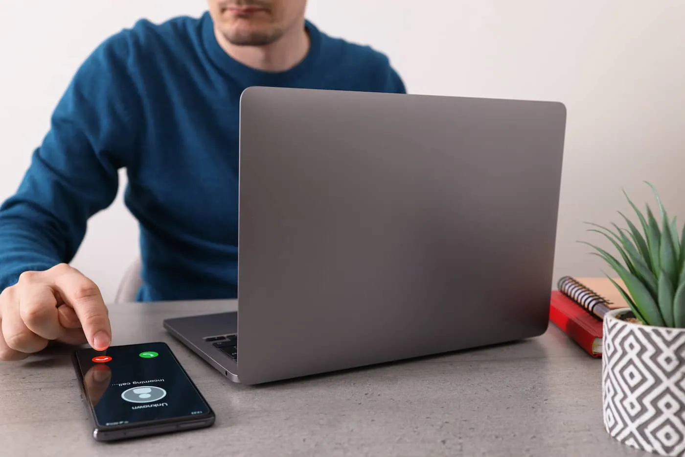 Partial view of a man in front of a laptop, while reaching to decline a call on his phone from an unknown number