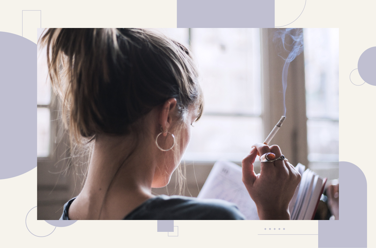 Woman with her hair up reading a book and smoking a cigarette inside