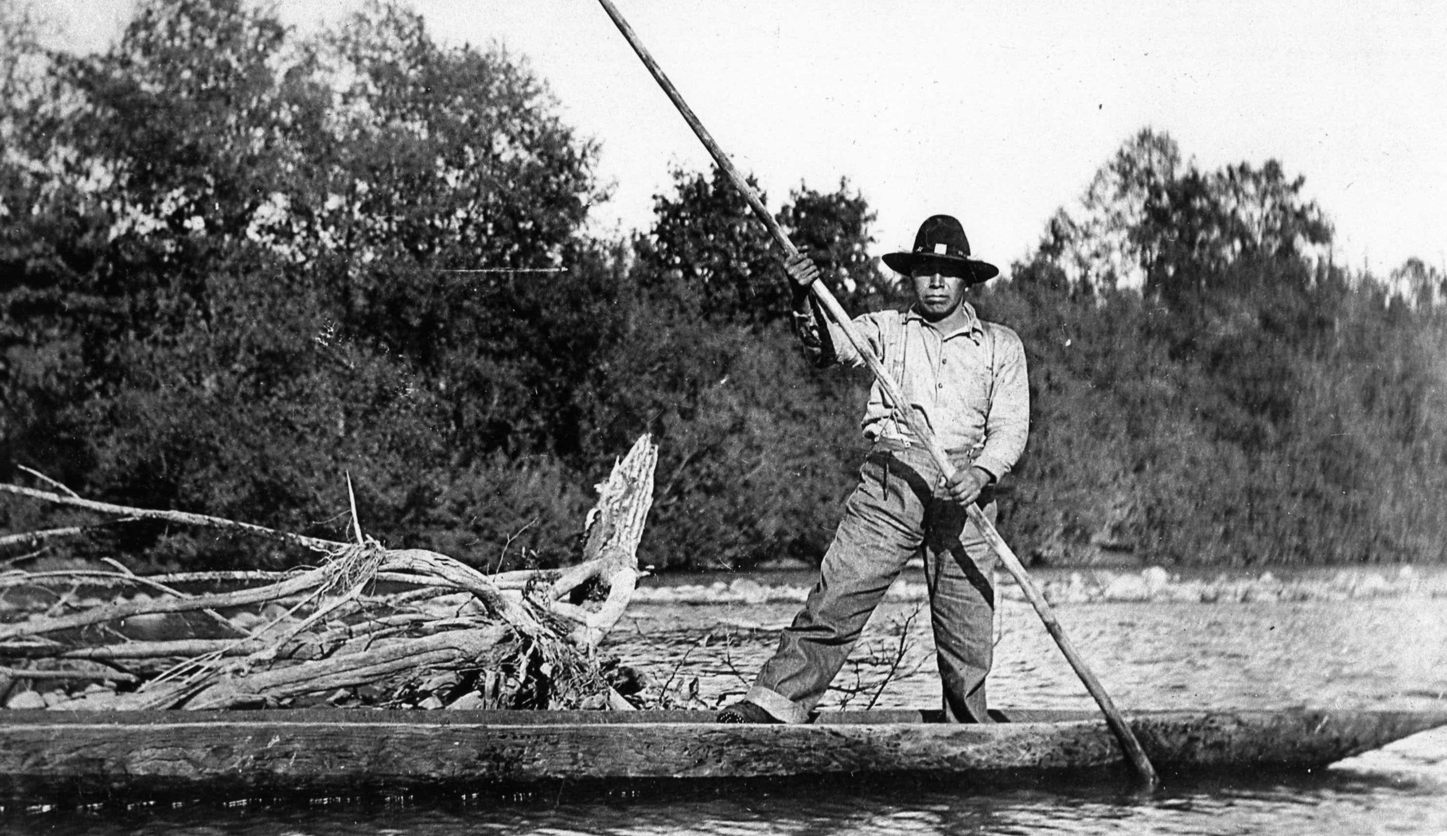 An aged, black and white photo of a Muckleshoot man standing on a wooden canoe on the water. He holds a long paddle in the water.