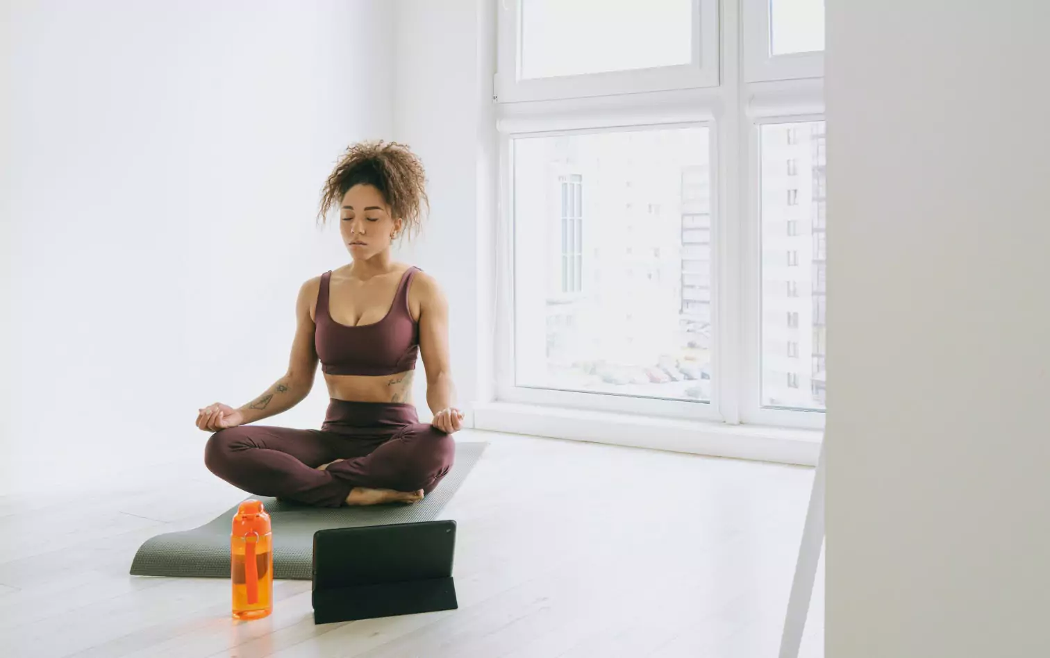 A woman on a yoga mat meditating with an orange bottle and ipad in front of her