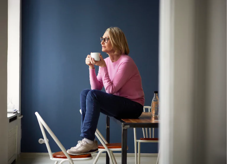 A person with mid-length blonde hair and glasses in a pink sweater and jeans sitting on a table holding a coffee cup