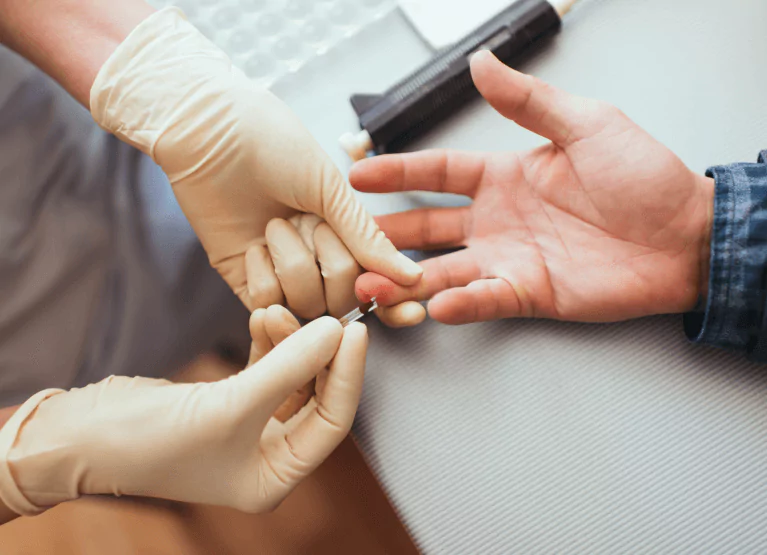 A medical professionals hands in white latex gloves administering a fingerprick glucose test to a hand placed on a table