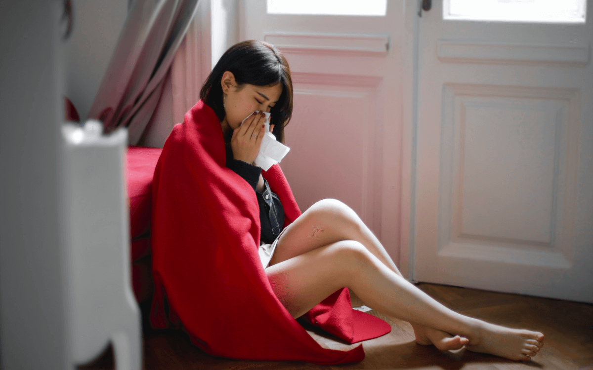 A woman with long dark hair sitting on the floor, wrapped in a red blanket and using a tissue
