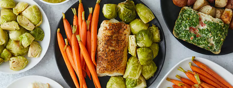 A close-up of a meal of protein, carrots and Brussels sprouts on a black plate, surrounded by other plates of similar food