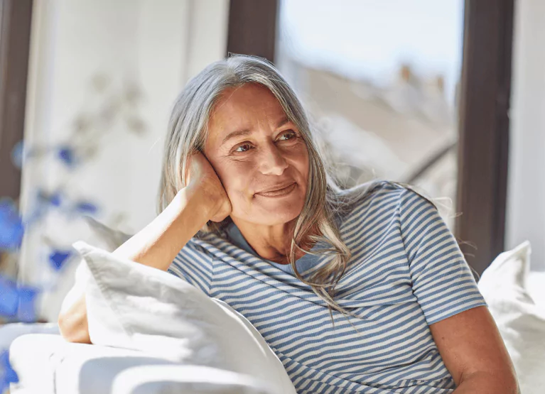 An older person with long grey hair in a striped t shirt leaning against cushions