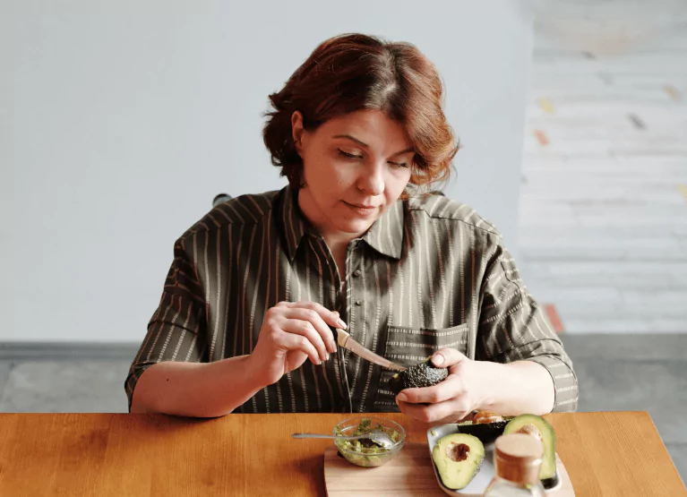 A person with mid-length brown hair sitting at a wooden table eating an avocado