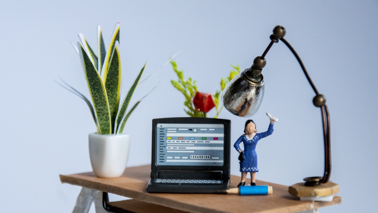 Ein Laptop auf einem Schreibtisch mit einer Tischlampe rechts daneben. Eine weibliche Office Miniaturfigur steht rechts neben dem Laptop.