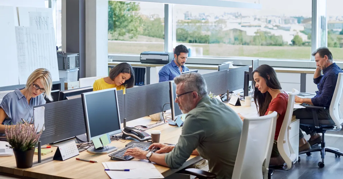 People working side to side on a large desk in an office on their computers.