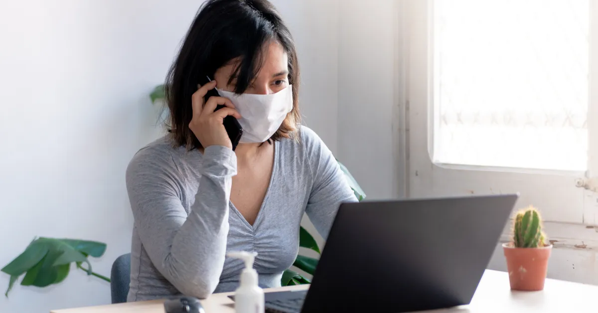 Women wearing mask talking on smartphone and working on laptop.