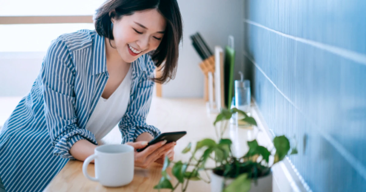 Smiling woman using smartphone and having a cup of coffee in the kitchen.