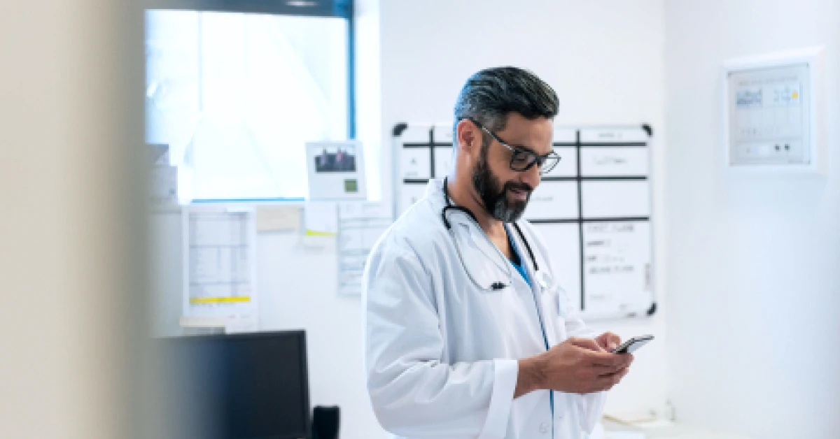 Smiling bearded doctor using smart phone in hospital.
