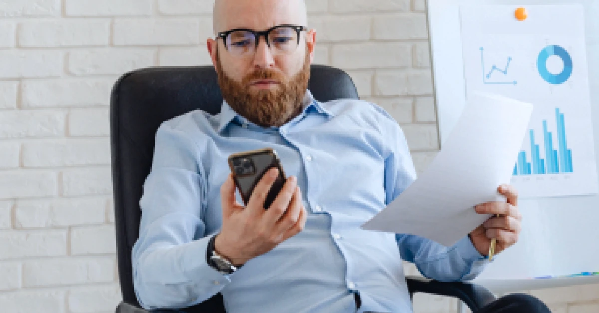 A businessman sitting on an office chair, holding a document and looking at his smartphone.
