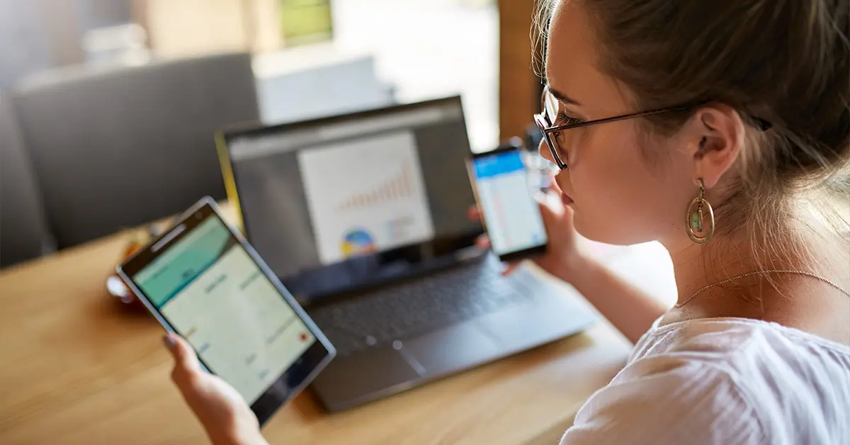 Woman looking at a tablet while holding a phone and working on her laptop.