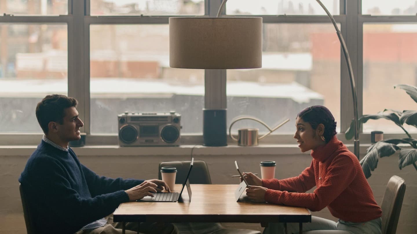 Two people with laptops speaking together and sitting at a table 