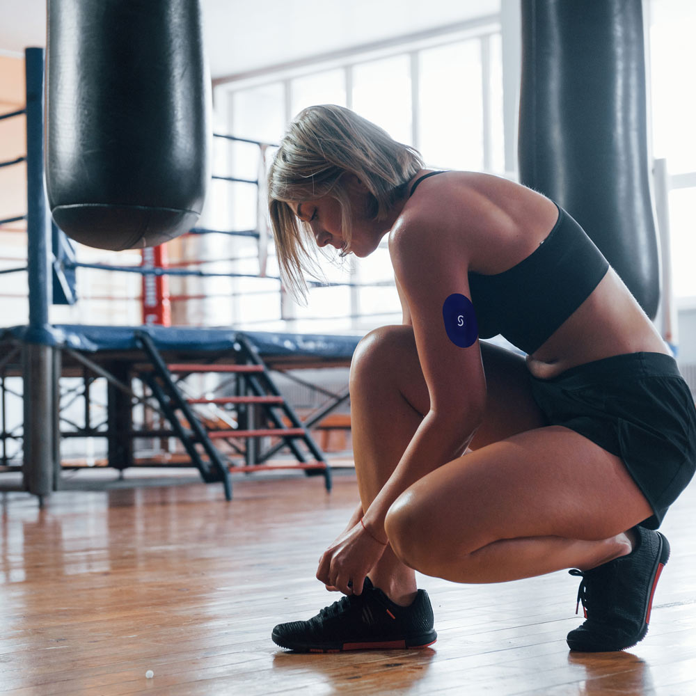 An older white woman crouching down, tying her shoe. She's in a gym and is wearing a grey sports bra and black shorts, and has a CGM and a Signos sports cover on the tricep of her left arm.