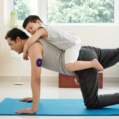 A boy is on his dad's back with his arms around his shoulders. The dad is on all fours, extending his right leg behind him, and is wearing a CGM with Signos sports cover on his left arm.