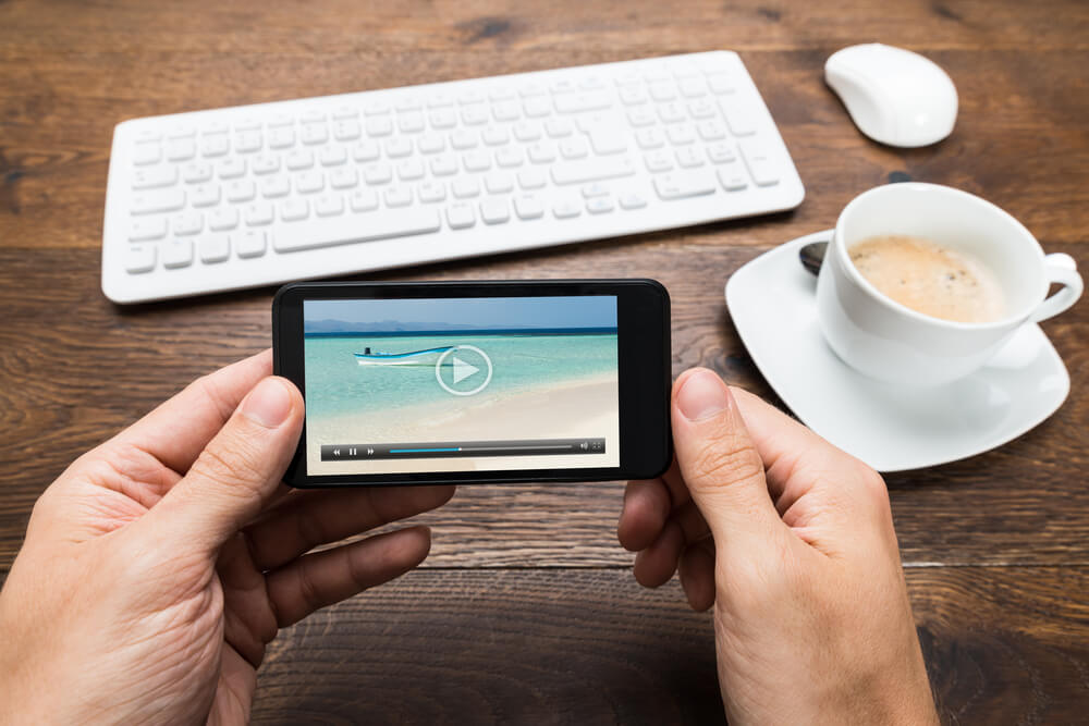 Close-up Of Person Watching Video On Mobile Phone With Cup Of Tea At Desk