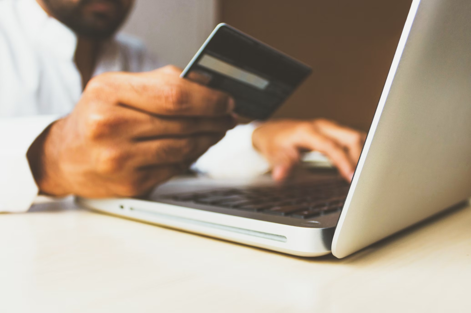 A photo of a man looking at the payment card in his hand while typing on a laptop keyboard