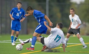 High school students playing soccer outside. Two of them wear blue uniforms and two wear white uniforms.